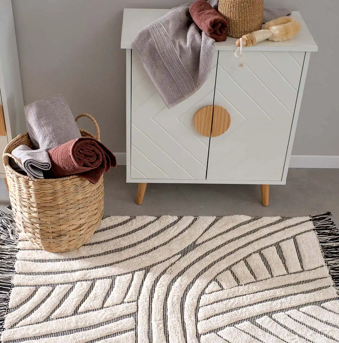 Badmat in lichte, neutrale kleuren met een geometrisch patroon, naast een rieten mand met handdoeken en een witte kast met houten handgrepen en opgevouwen handdoeken op het blad.