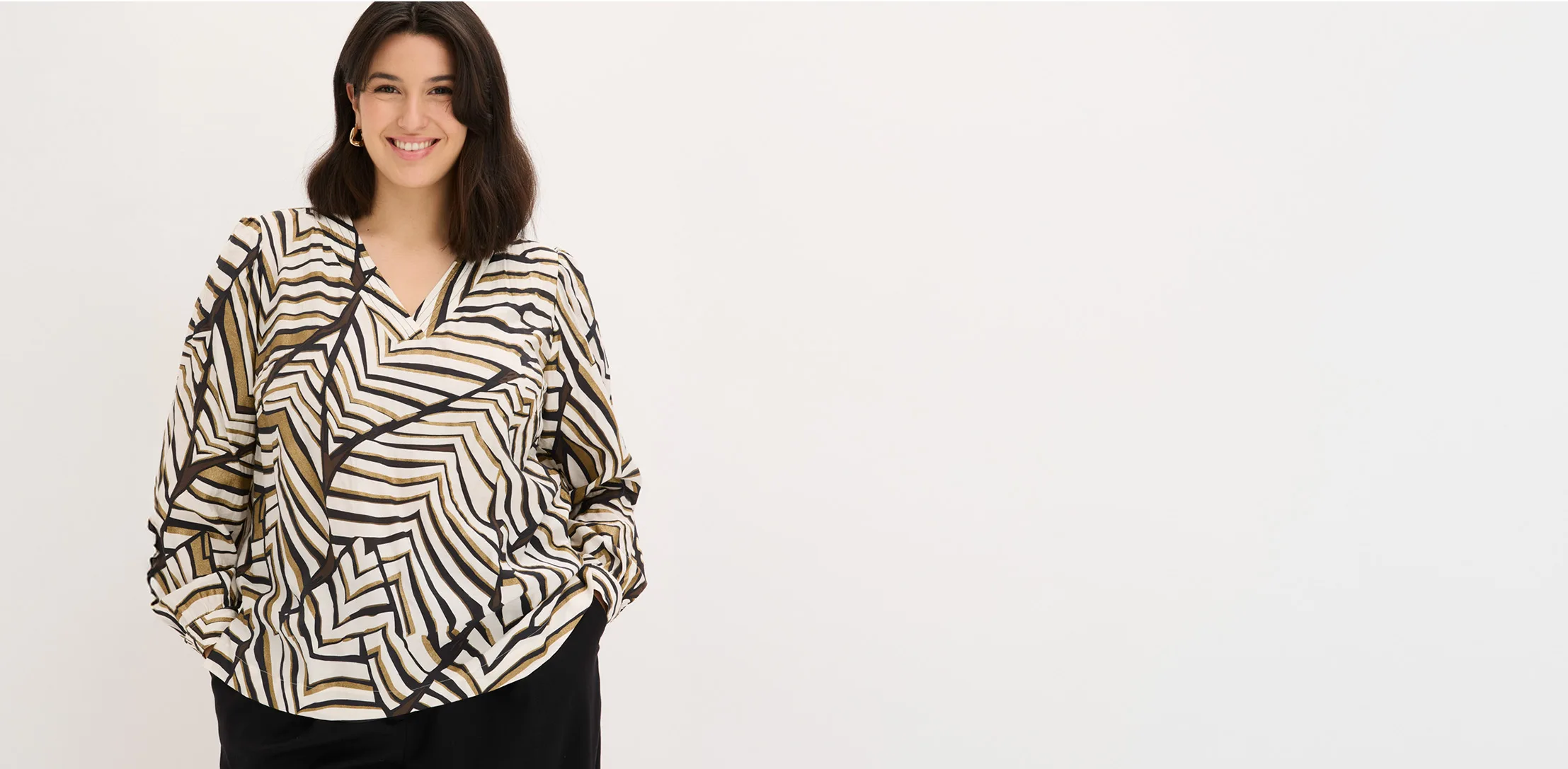 Het model draagt een ruime blouse met lange mouwen in tinten beige, zwart en wit, met een golvend geometrisch patroon.