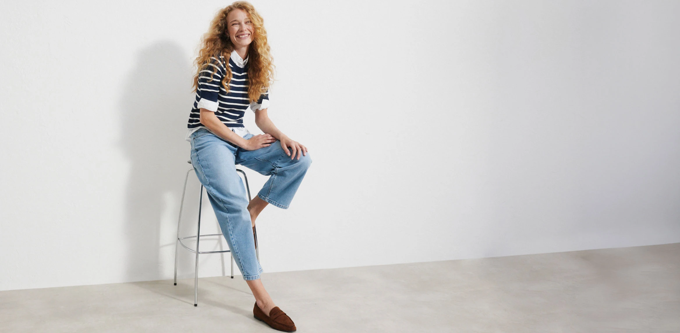 "Donkerblauwe barrel‑jeans met een hoge taille en een afgeronde, taps toelopende pijp, gemaakt van glad denim, gecombineerd met een witte trui met V‑hals en contrasterende details en bruine enkellaarsjes met een lage hak."