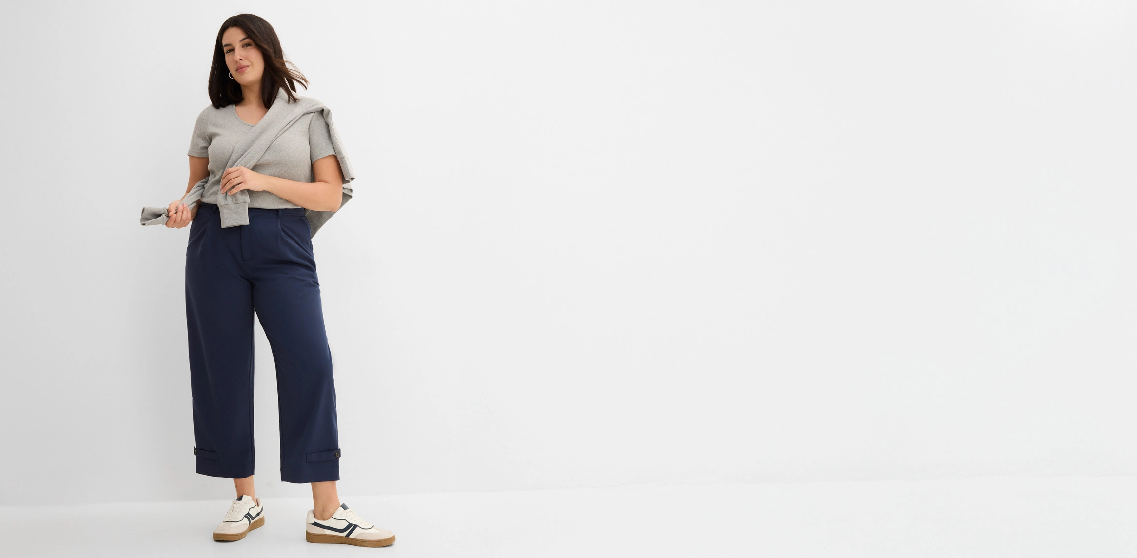 Grijs T-shirt gecombineerd met een donkerblauwe broek met een decoratief paneel in de taille, een vest en witte sneakers.