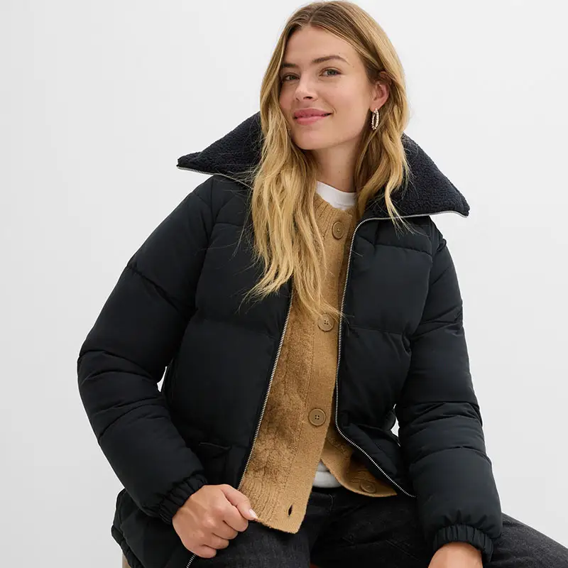 Zwarte winterjas met hoge kraag over beige gebreide trui en wit shirt.