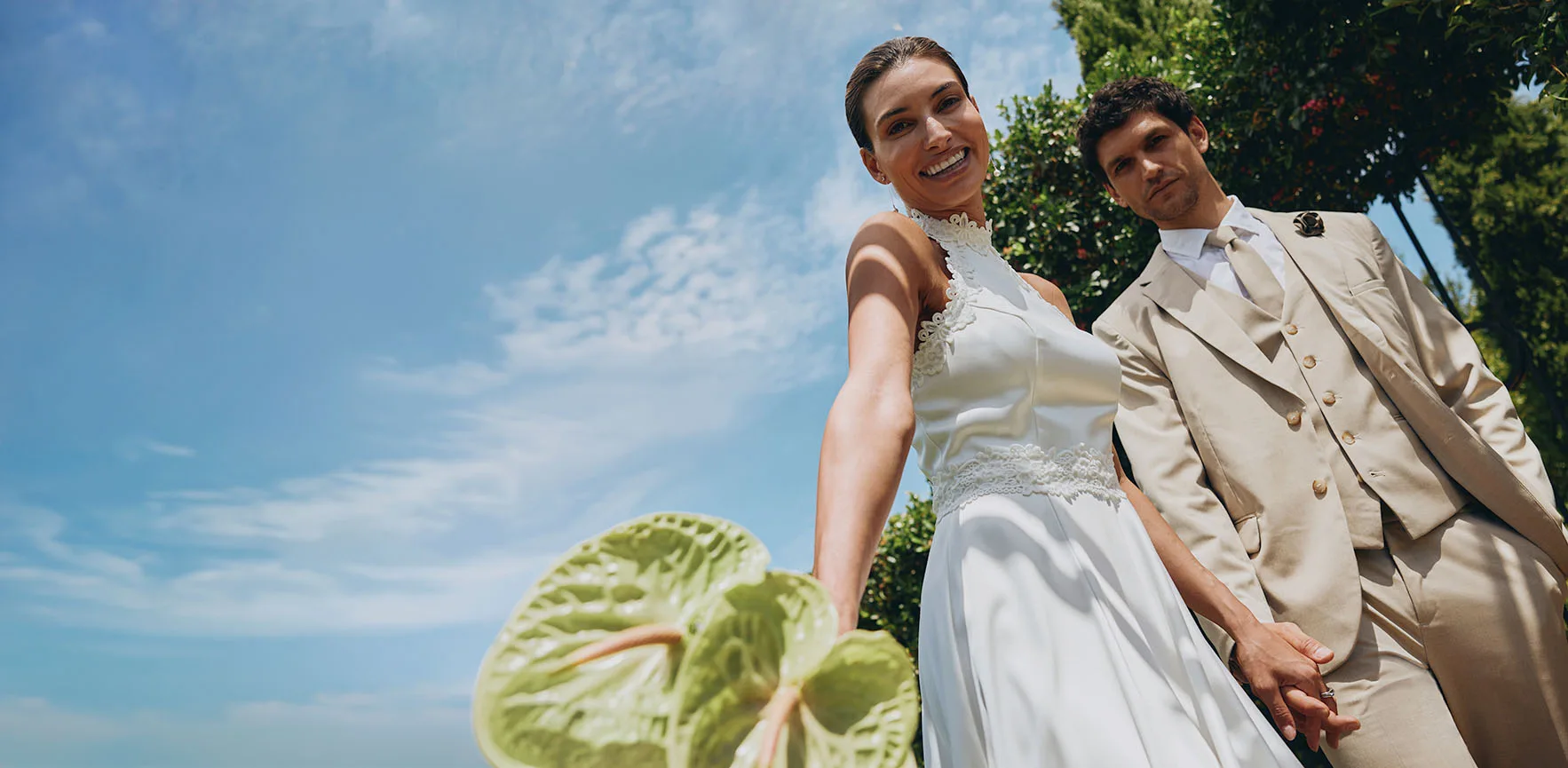 Elegant bruidspaar in de zon: zij in een witte jurk en hij in een beige pak met zonnebril.