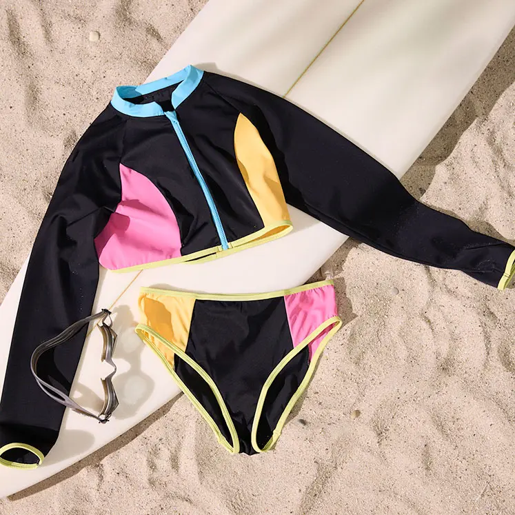 Kleurrijk badpak met lange mouwen en een bikini op het zand naast een surfplank.