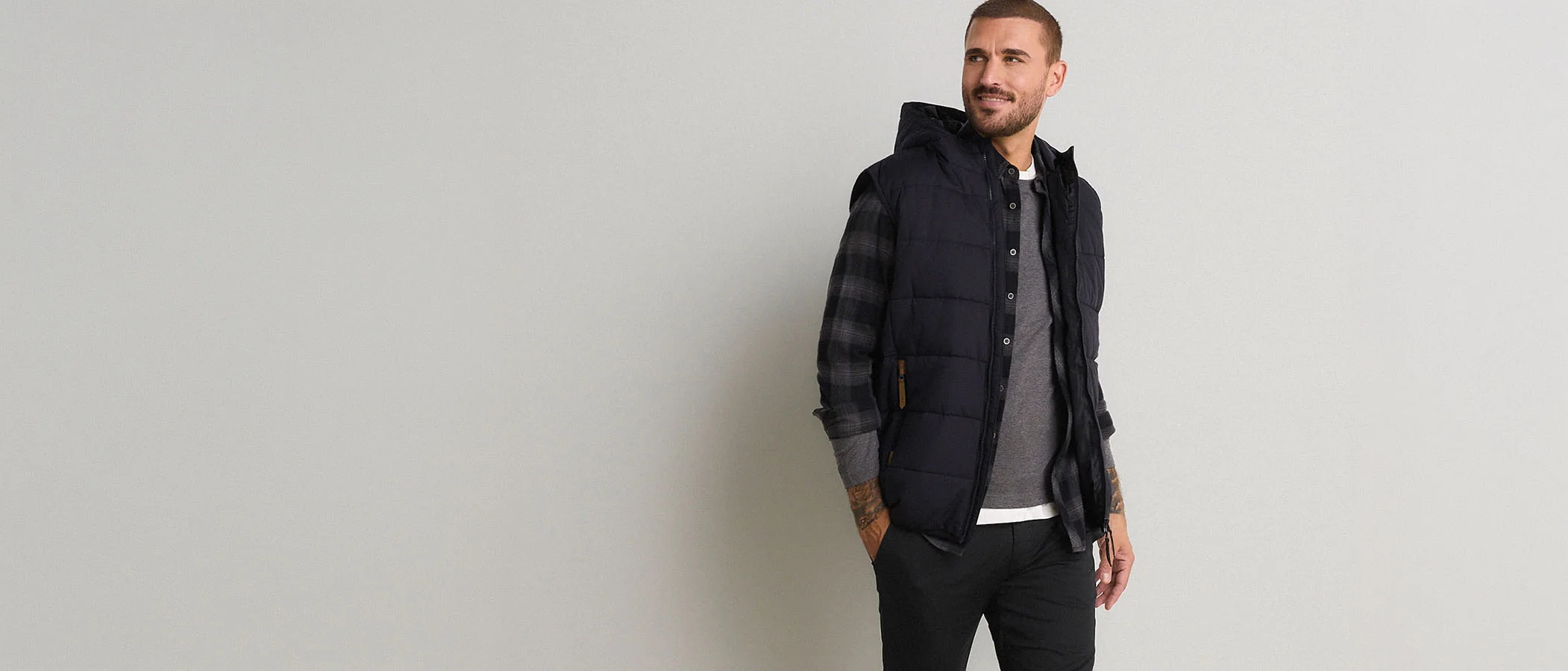Donkere gewatteerde bodywarmer met capuchon, geruit overhemd, grijs shirt en zwarte broek.