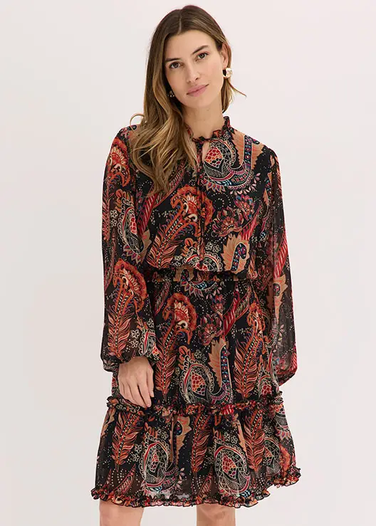 Ze draagt een soepelvallende jurk met kleurrijk paisley dessin en ruchedetails.