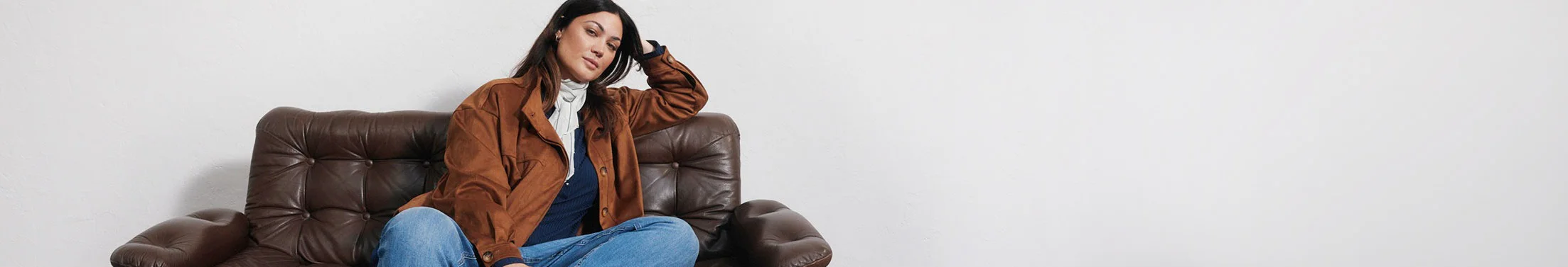 Jas in suèdelook, blauwe jeans, donker shirt, licht sjaaltje en laarzen.