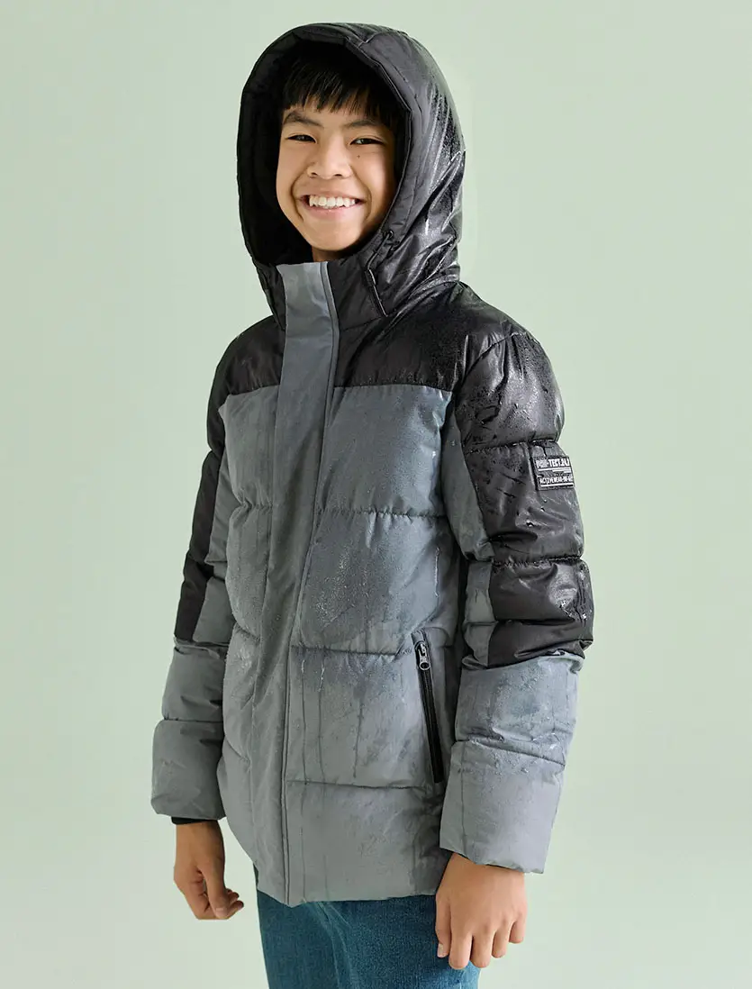 Deze jongen draagt een stylish tweekleurige gewatteerde jas met capuchon en een jeans.