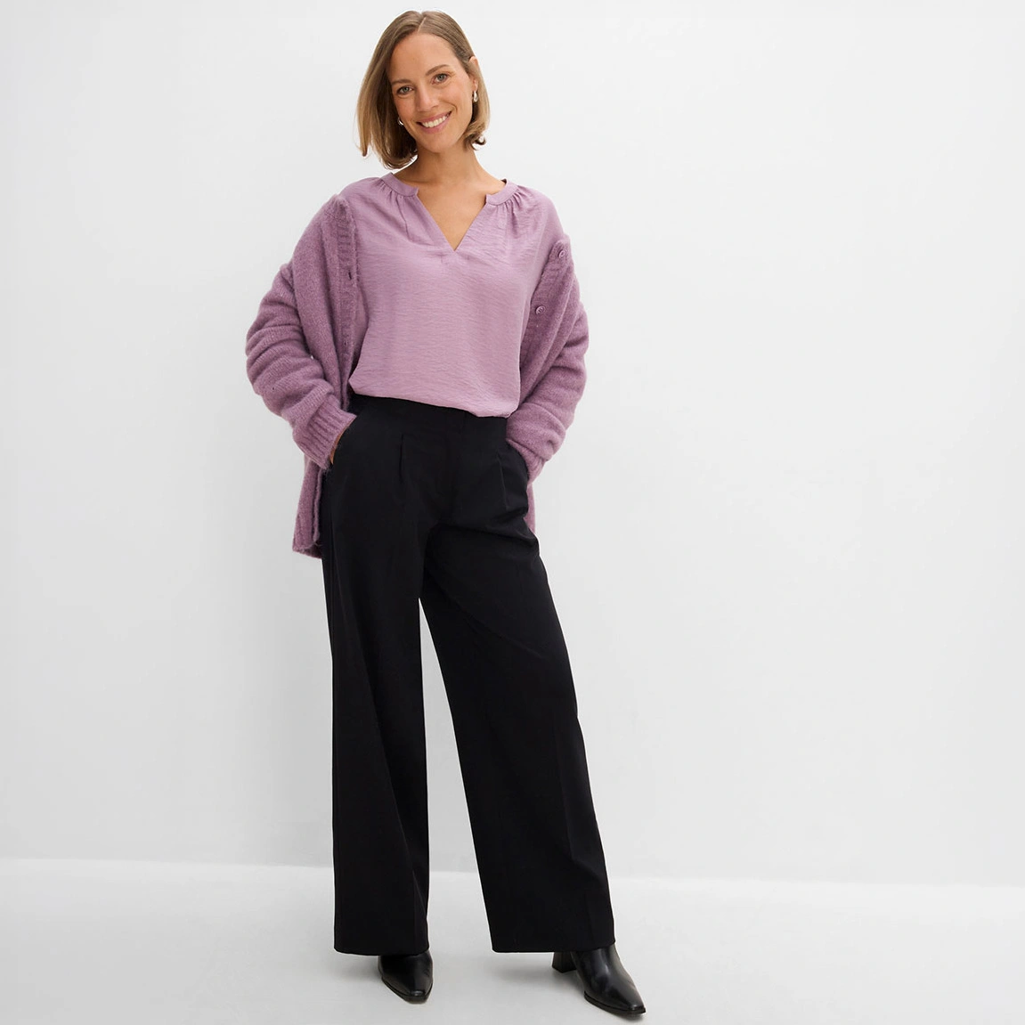 Ze draagt een lila blouse met een gebreid vest en een wijde zwarte broek.
