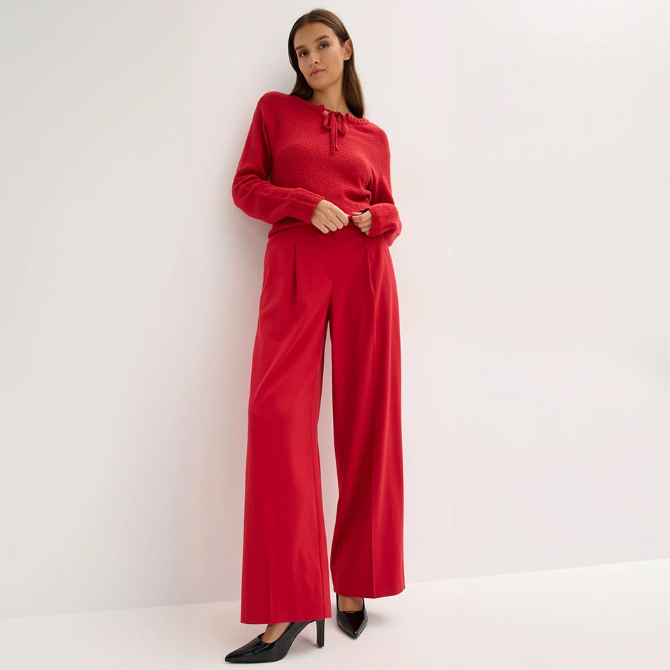Ze draagt een lila blouse met een gebreid vest en een wijde zwarte broek.