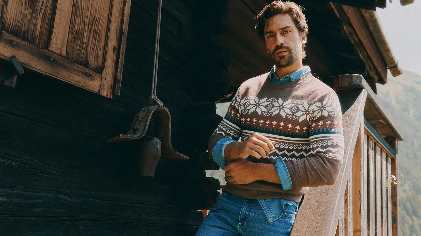 Hij draagt een bruine trui met motief, een spijkeroverhemd en een blauwe jeans.