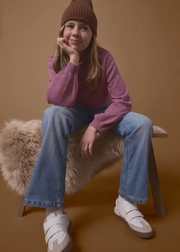 Dit meisje draagt een bruine beanie, een pink trui, een blauwe jeans en witte sneakers.