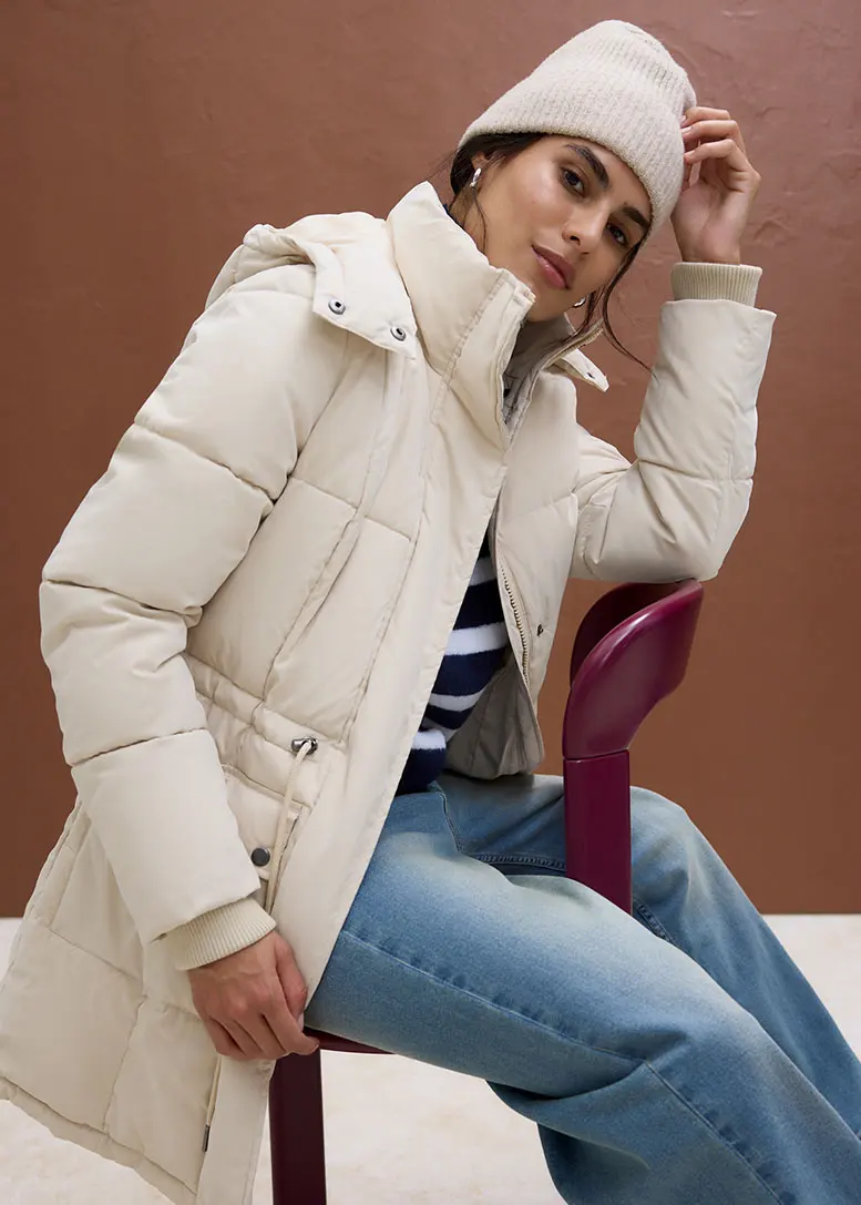 Deze vrouw draagt een beige gewatteerde jas, een gestreept shirt, een jeans en een muts.