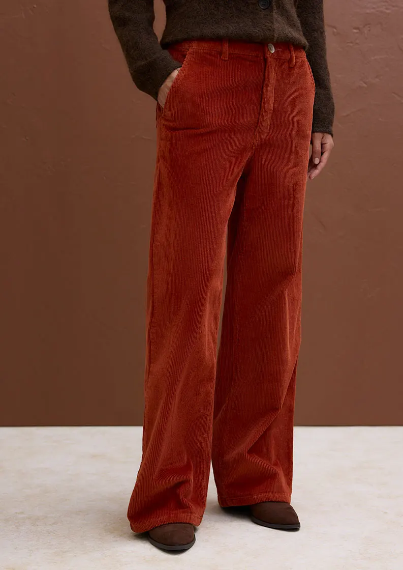 Zij draagt een gebreid vest met motief over een witte overhemdblouse met een oranje corduroy broek eronder.